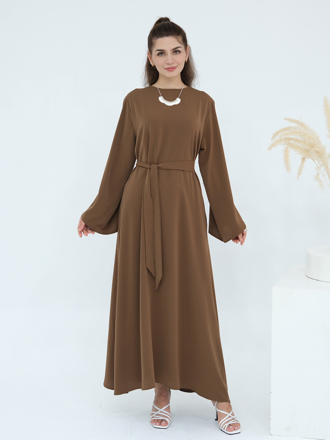 A woman modeling a long-sleeved brown maxi dress with a matching waist tie belt, standing against a clean white studio background.