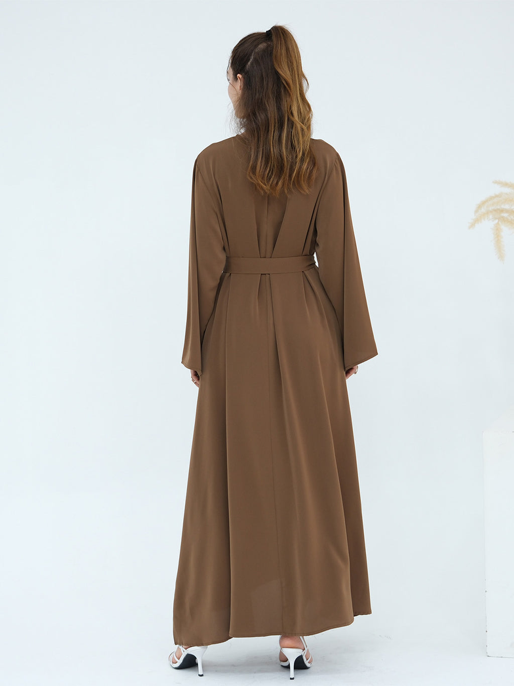 A woman viewed from behind wearing a long brown abaya dress with long sleeves, a matching waist tie belt, and white high heels, set against a clean white studio background.