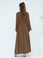 A woman viewed from behind wearing a long brown abaya dress with long sleeves, a matching waist tie belt, and white high heels, set against a clean white studio background.