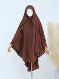 A brown modest garment set displayed on a mannequin, featuring a draped hooded outer layer with wide sleeves over a matching inner top. The mannequin is shown against a light studio background with decorative pampas grass on the right.