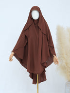 A brown modest garment set displayed on a mannequin, featuring a draped hooded outer layer with wide sleeves over a matching inner top. The mannequin is shown against a light studio background with decorative pampas grass on the right.