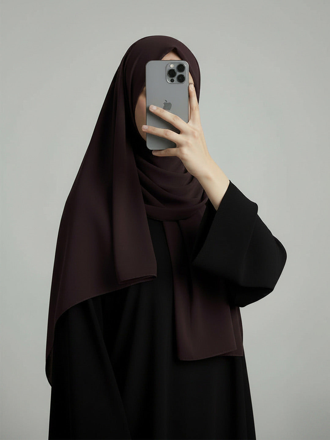 A woman wearing a dark brown hijab and a black abaya takes a mirror selfie with a gray smartphone against a plain light gray background.