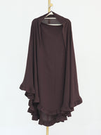 A dark brown draped tunic top with wide sleeves and layered ruffles along the hem, hanging on a wooden hanger against a plain white background.