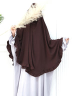 A woman wearing a dark brown full-body modest garment with wide flutter sleeves and ruffled edges, posed against a white paneled background while holding a dried pampas grass stem.