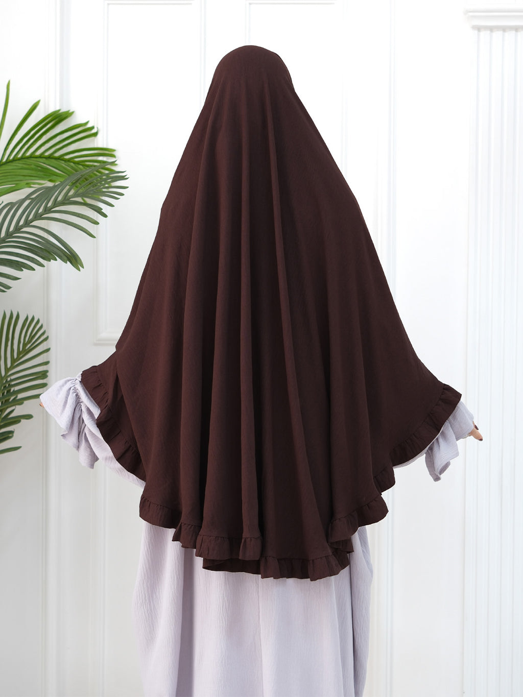 A person shown from the back wearing a dark brown full-body modest garment or hijab with wide draped sleeves over a light lavender dress, standing against a white paneled wall with a green plant leaf visible on the left.