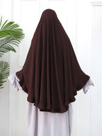 A person shown from the back wearing a dark brown full-body modest garment or hijab with wide draped sleeves over a light lavender dress, standing against a white paneled wall with a green plant leaf visible on the left.