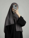 A woman wearing a dark gray hijab and a black abaya takes a mirror selfie with her phone against a solid gray background.