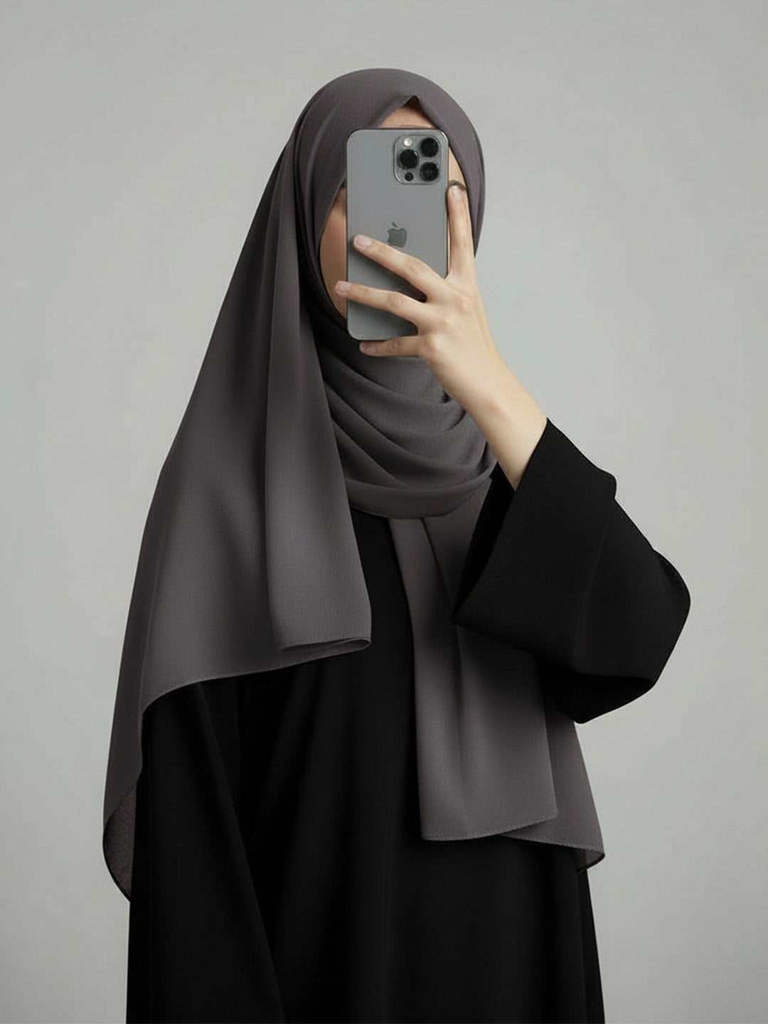 A woman wearing a dark gray hijab and a black abaya takes a mirror selfie with her phone against a solid gray background.
