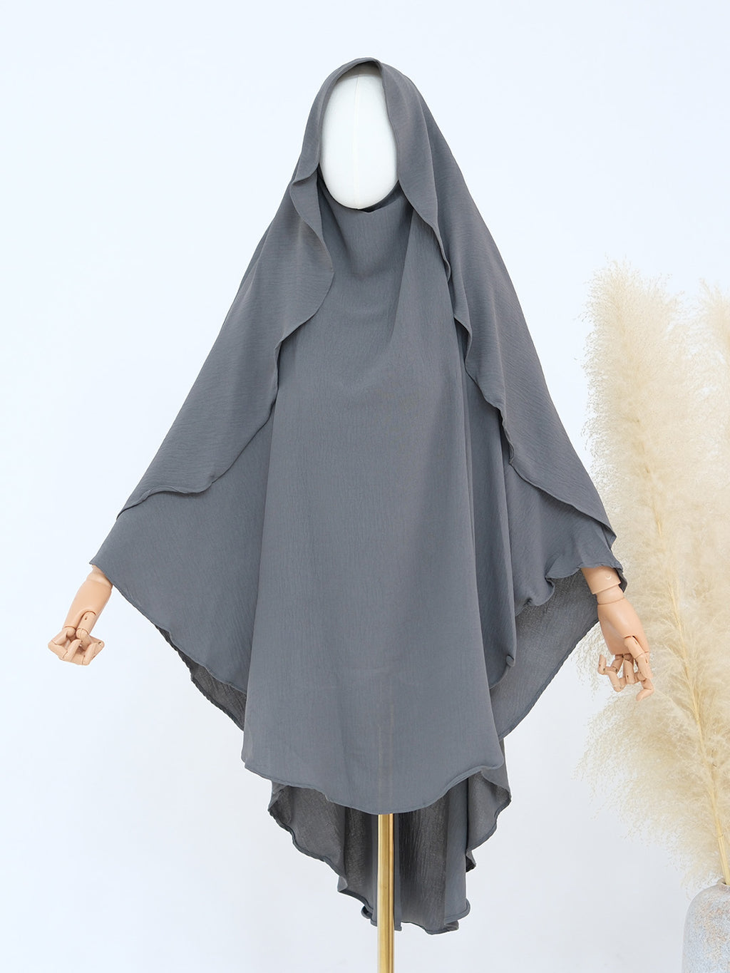 A dark gray modest hooded garment displayed on a mannequin with wooden arms, set against a light background with a decorative pampas grass plant on the right.