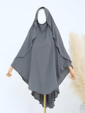A dark gray modest hooded garment displayed on a mannequin with wooden arms, set against a light background with a decorative pampas grass plant on the right.