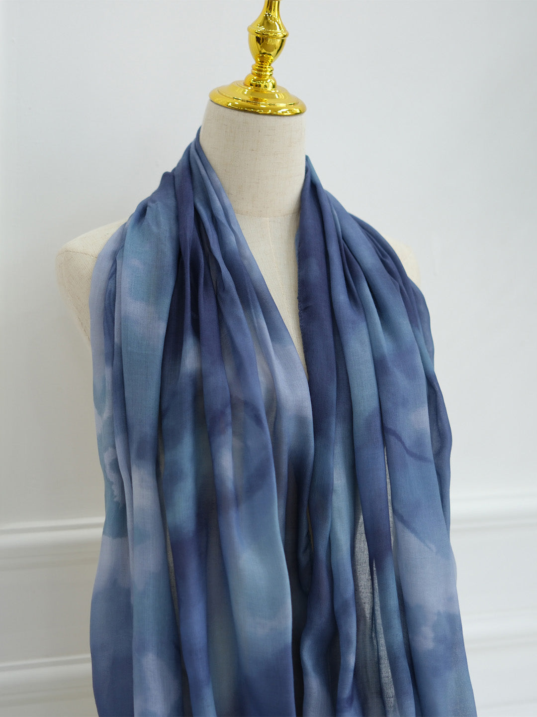 A lightweight blue and navy tie-dye scarf draped on a cream-colored dress form with a gold finial, set against a plain white wall.