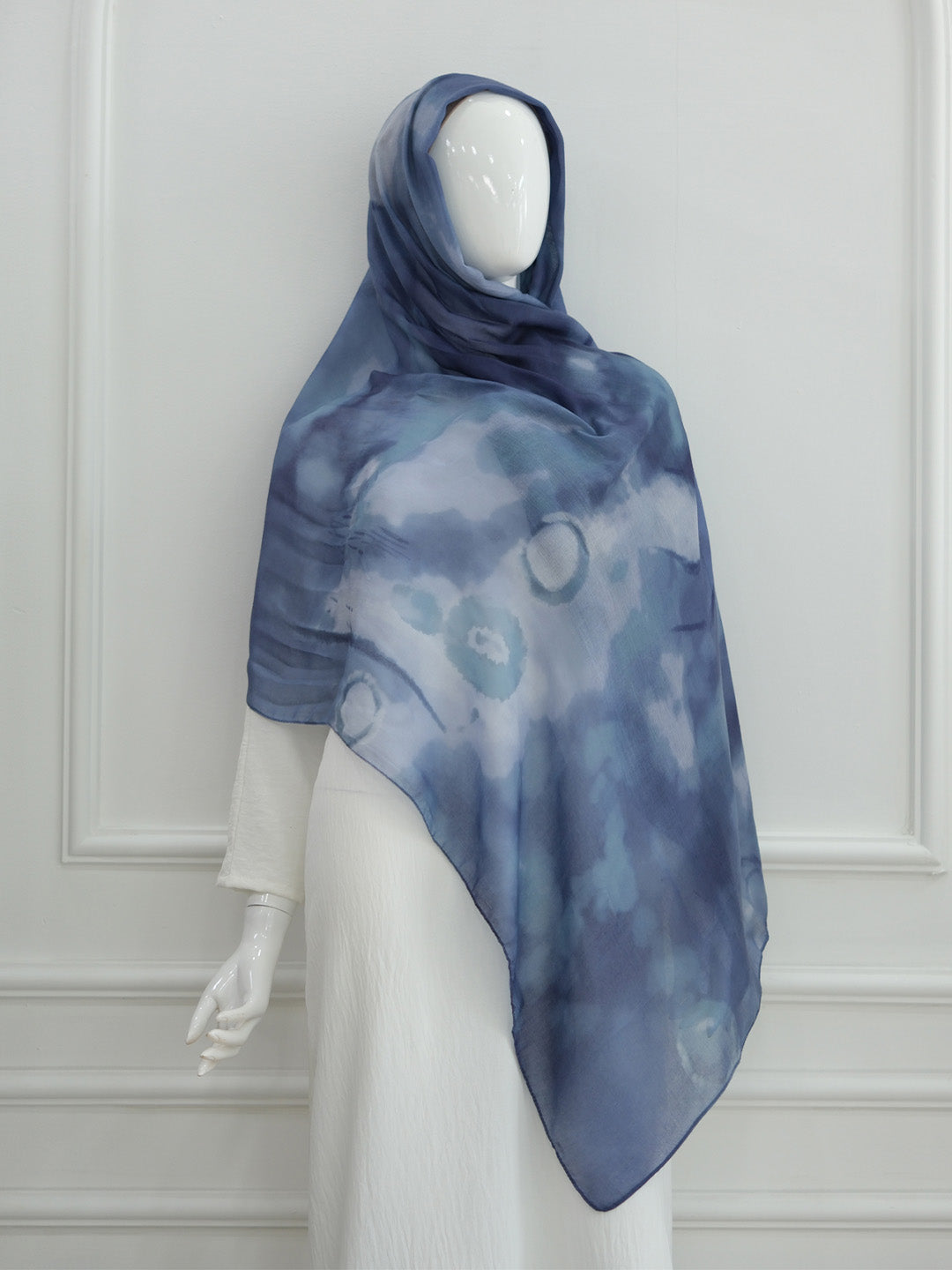 A blue and gray patterned hijab scarf draped on a white mannequin against a white paneled wall.