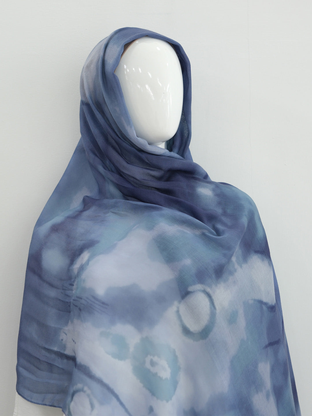 A blue and white tie-dye patterned hijab draped on a white mannequin head, set against a plain light gray background.