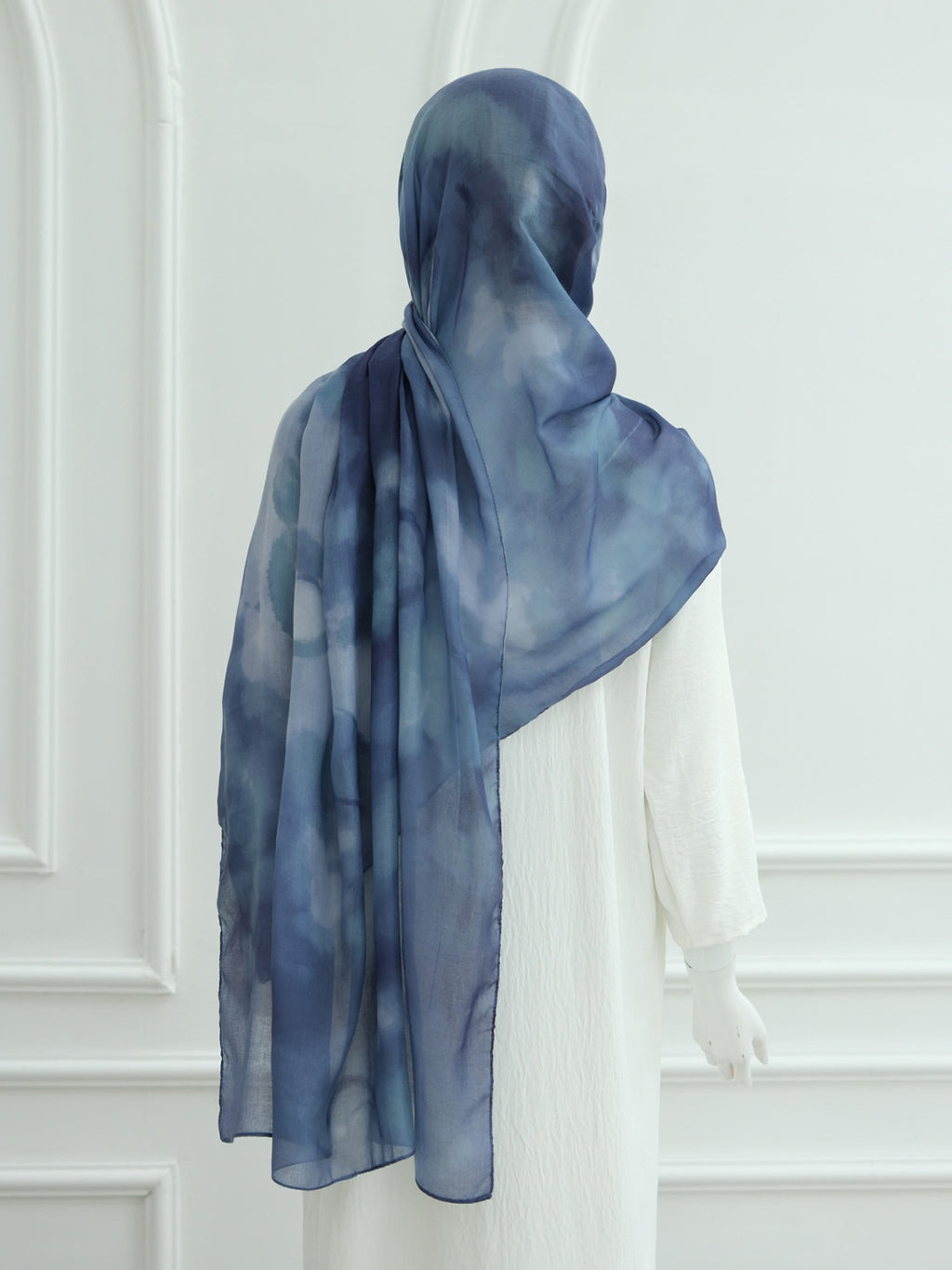 A blue and navy watercolor-patterned hijab draped on a white mannequin, shown from the back against a white paneled wall.