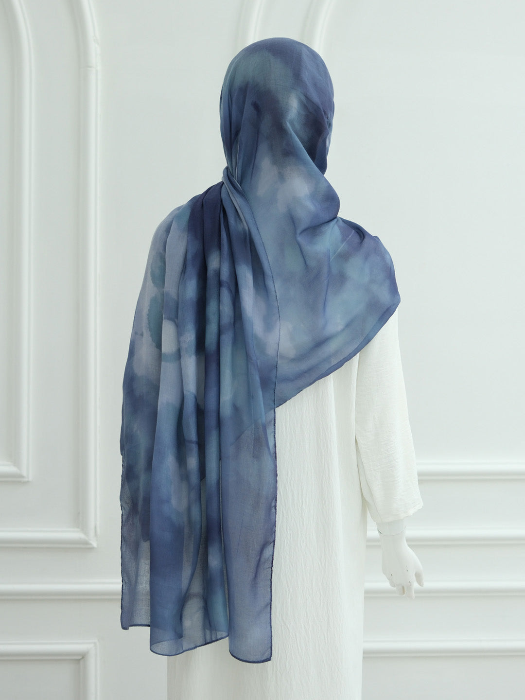 A blue and navy watercolor-patterned hijab draped on a white mannequin, shown from the back against a white paneled wall.