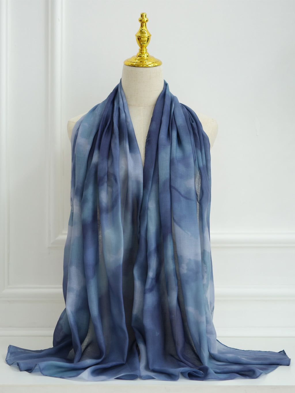 A lightweight blue scarf with a soft tie-dye or watercolor pattern, draped over a cream-colored mannequin torso with a gold finial, set against a white paneled wall.