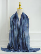 A lightweight blue scarf with a soft tie-dye or watercolor pattern, draped over a cream-colored mannequin torso with a gold finial, set against a white paneled wall.