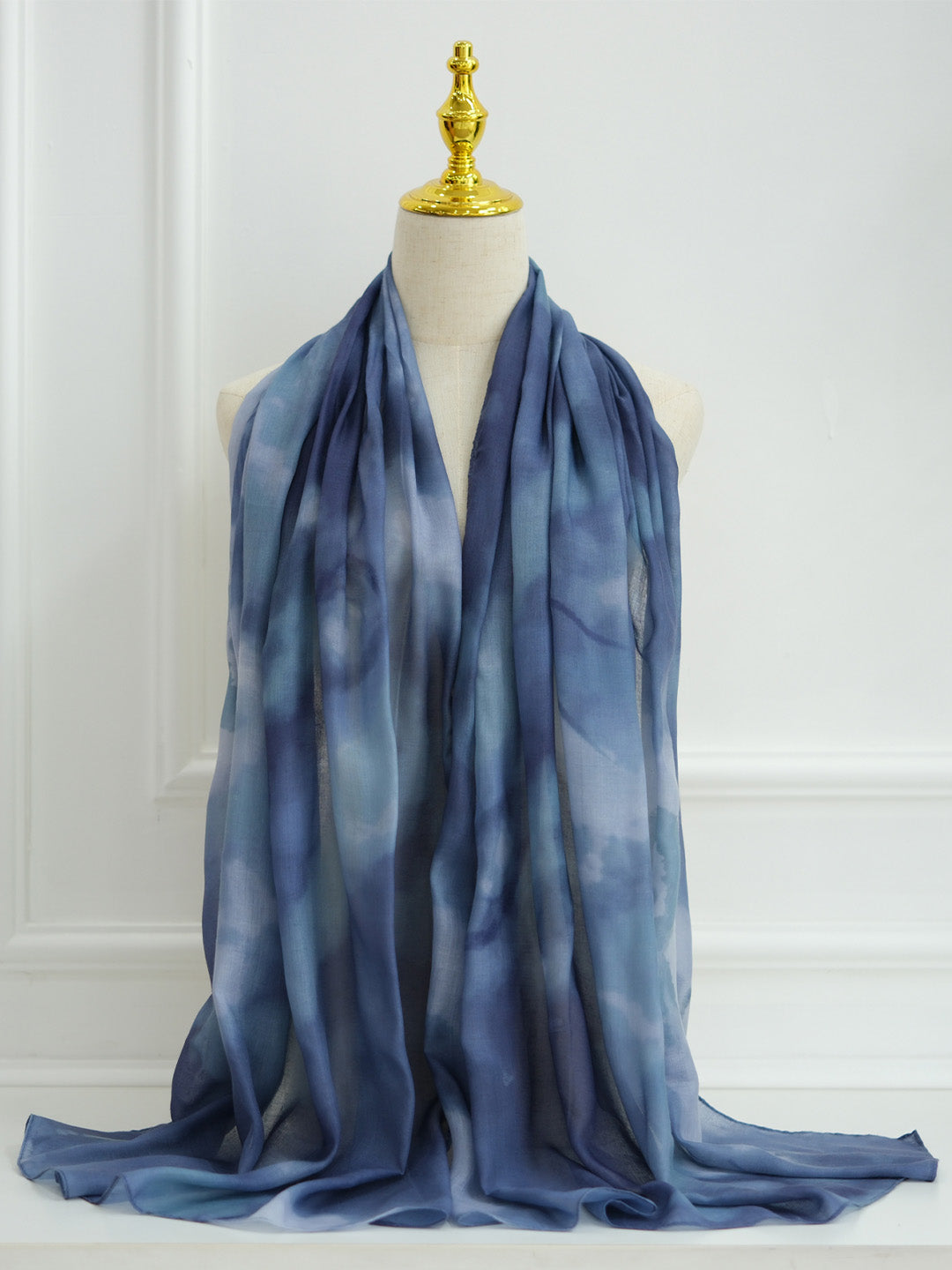 A lightweight blue scarf with a soft tie-dye or watercolor pattern, draped over a cream-colored mannequin torso with a gold finial, set against a white paneled wall.