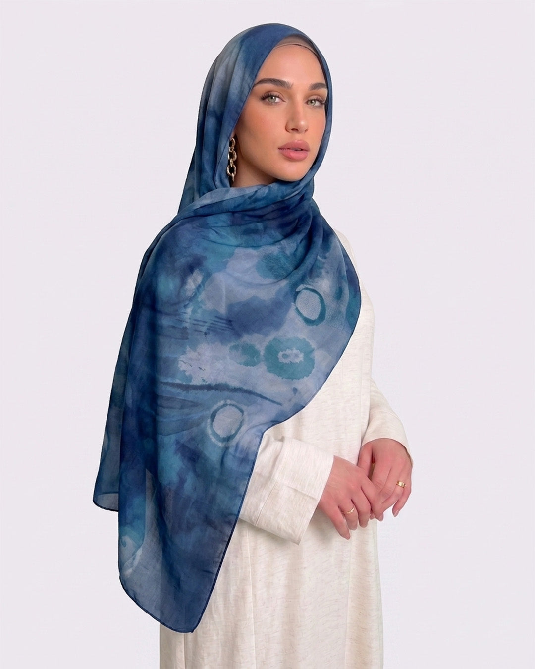 A woman wearing an artistic printed modal hijab with a watercolor shibori pattern in shades of indigo blue, denim, and teal, paired with a cream outfit.