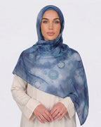 A woman wearing an artistic printed modal hijab with a watercolor shibori pattern in shades of indigo blue, denim, and teal, paired with a cream outfit.
