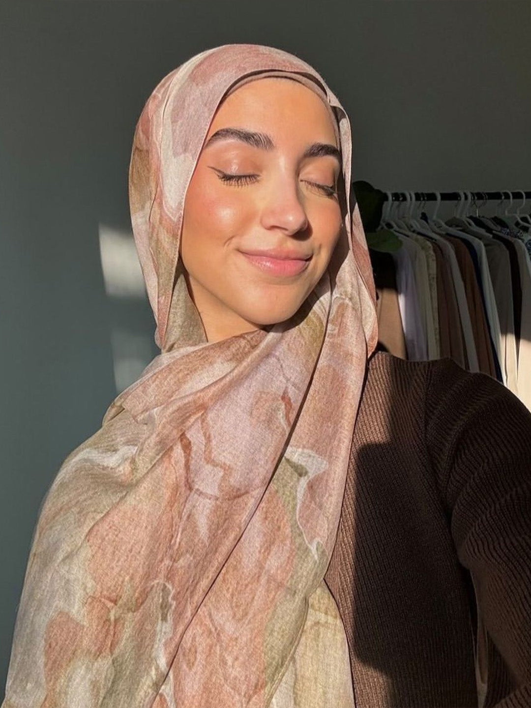 A woman wearing a light pink and beige patterned hijab with a brown ribbed top, captured in a warm, sunlit indoor setting with a clothing rack in the background.