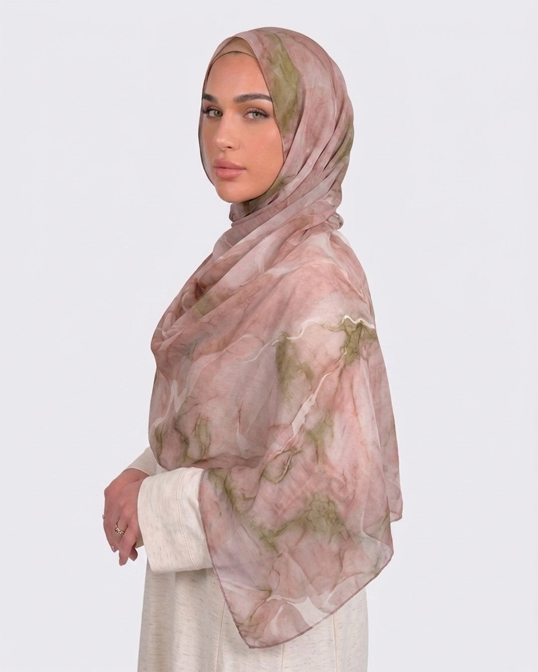A woman wearing a soft dusty rose and blush pink printed modal hijab with fine marble veins of sage and olive green, paired with a cream outfit.