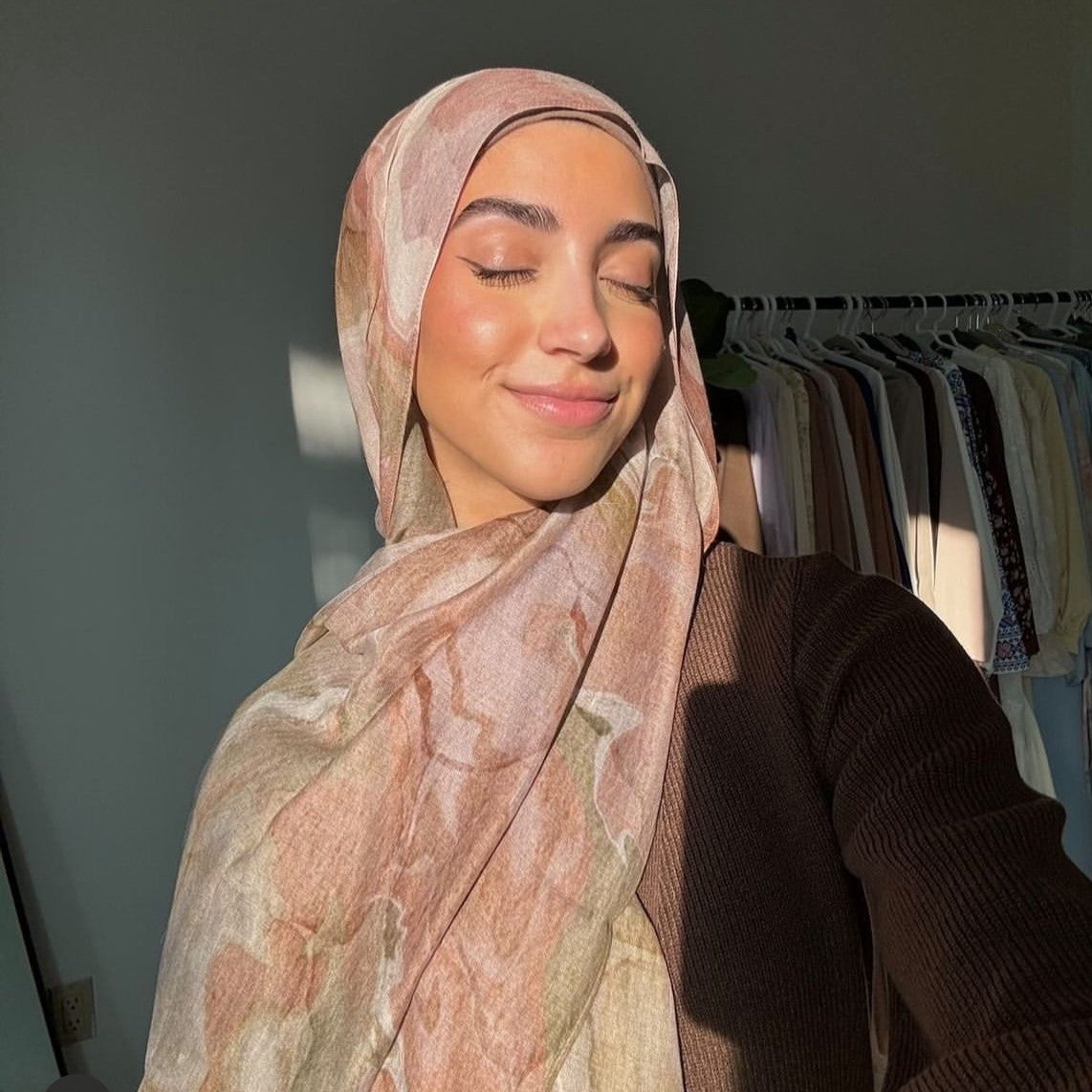 Woman wearing a patterned hijab in front of a clothing rack.