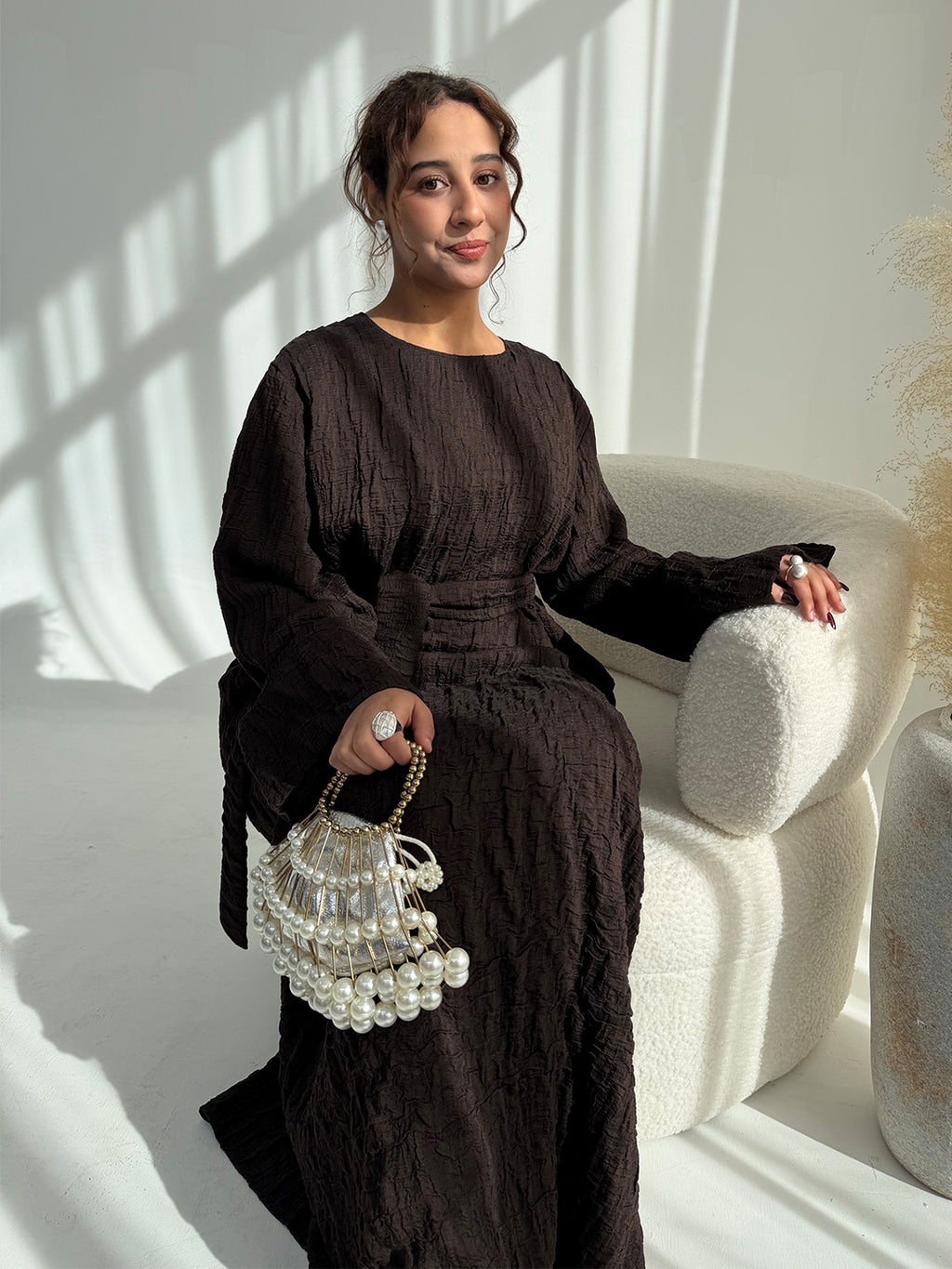 A woman wearing a dark brown textured long-sleeve maxi dress sits on a cream-colored armchair, holding a pearl-beaded handbag.