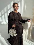 A woman wearing a dark brown textured long-sleeve maxi dress sits on a cream-colored armchair, holding a pearl-beaded handbag.