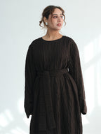 A woman wearing a dark brown crinkled cotton dress with long sleeves and a matching fabric belt, standing against a light gray background with soft window shadows.