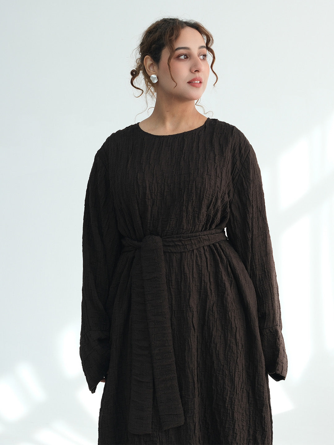 A woman wearing a dark brown crinkled cotton dress with long sleeves and a matching fabric belt, standing against a light gray background with soft window shadows.