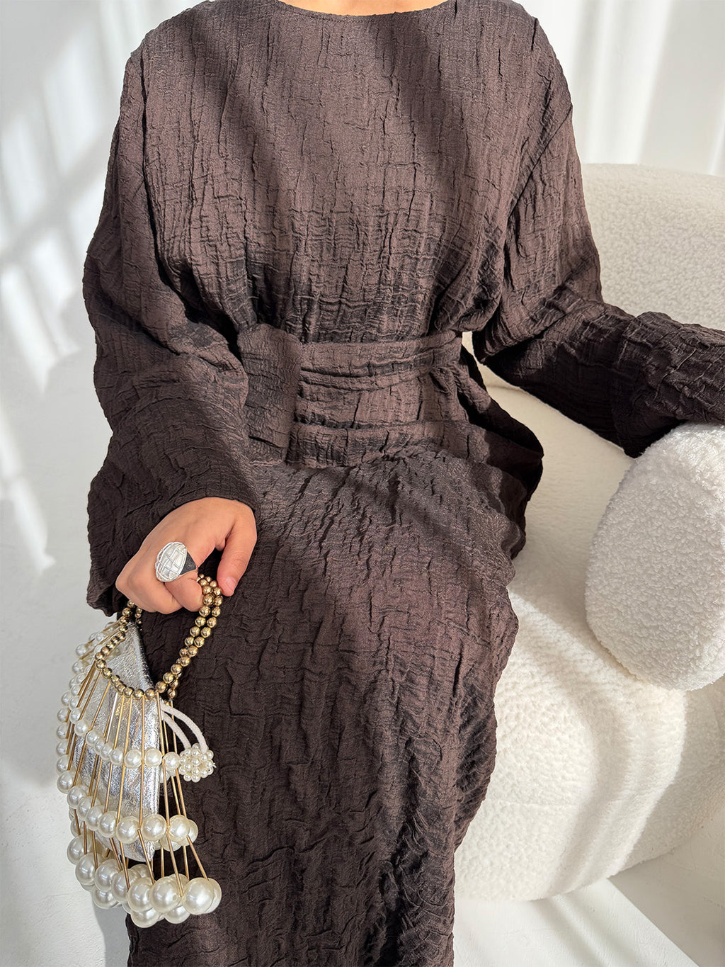 A person seated on a cream textured chair wearing a dark brown crinkled long-sleeve dress with a self-tie waist belt. The person is holding a pearl-bead fringe handbag with a gold chain strap and wearing a large silver ring.