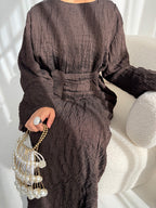 A person seated on a cream textured chair wearing a dark brown crinkled long-sleeve dress with a self-tie waist belt. The person is holding a pearl-bead fringe handbag with a gold chain strap and wearing a large silver ring.
