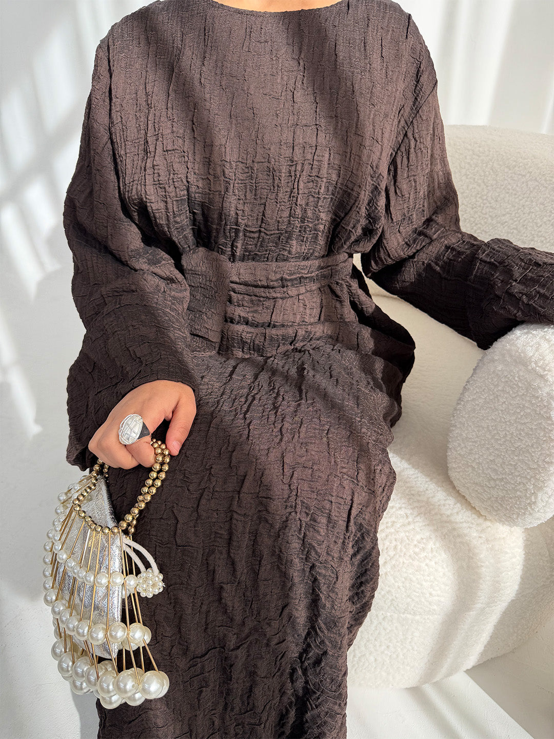 A person seated on a cream textured chair wearing a dark brown crinkled long-sleeve dress with a self-tie waist belt. The person is holding a pearl-bead fringe handbag with a gold chain strap and wearing a large silver ring.