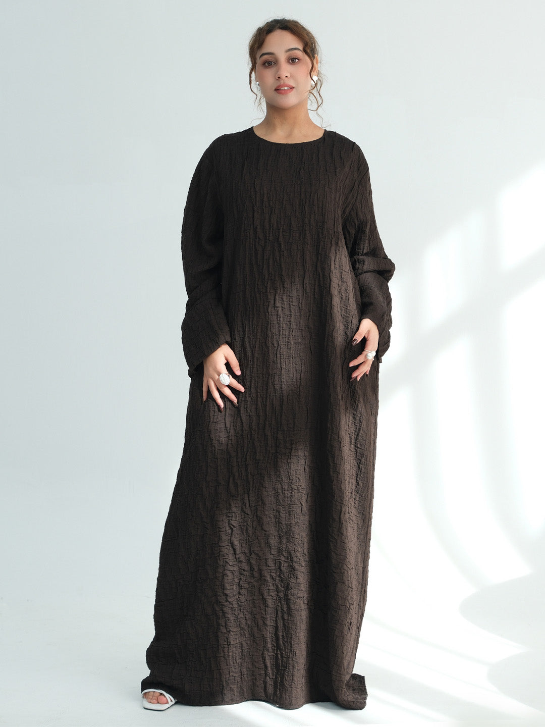 A woman modeling a long-sleeved, floor-length dark brown textured dress against a light gray studio background with soft window-like shadows.