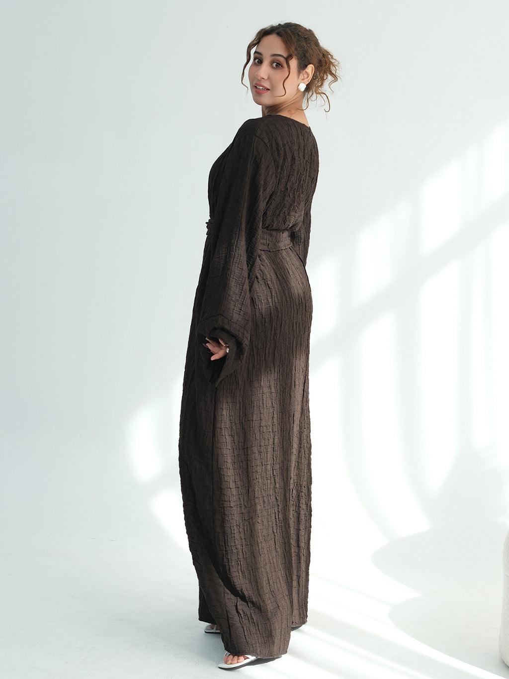 A woman wearing a dark brown textured maxi dress with long sleeves and a side pocket, standing in a bright white studio with soft window-like shadows.