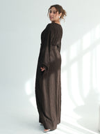 A woman wearing a dark brown textured maxi dress with long sleeves and a side pocket, standing in a bright white studio with soft window-like shadows.