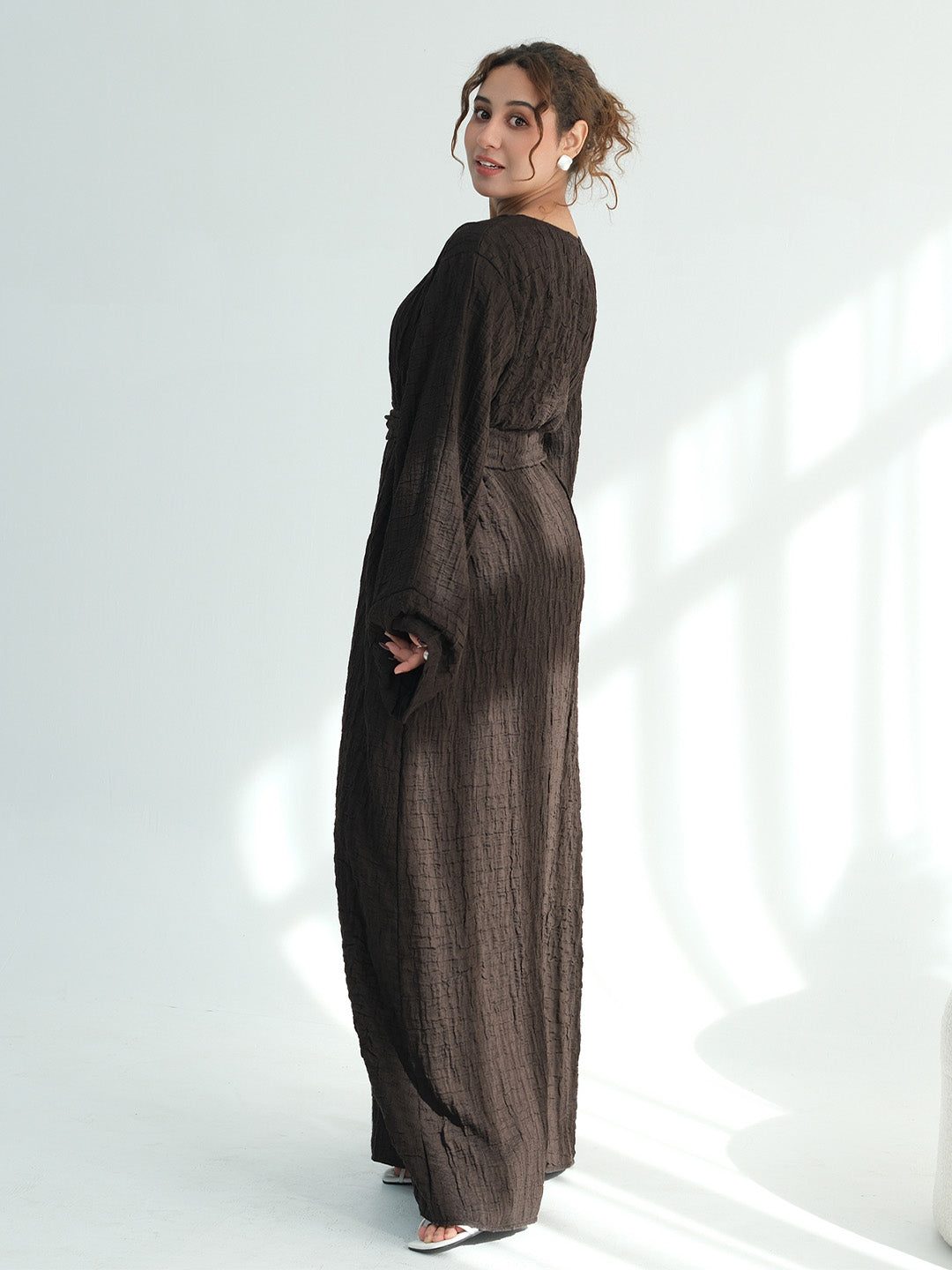 A woman wearing a dark brown textured maxi dress with long sleeves and a side pocket, standing in a bright white studio with soft window-like shadows.