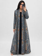 A woman takes a mirror selfie wearing a long, open-front, floor-length kaftan dress with a detailed blue, gray, and brown geometric and floral pattern over a black dress, set against a plain light gray background.