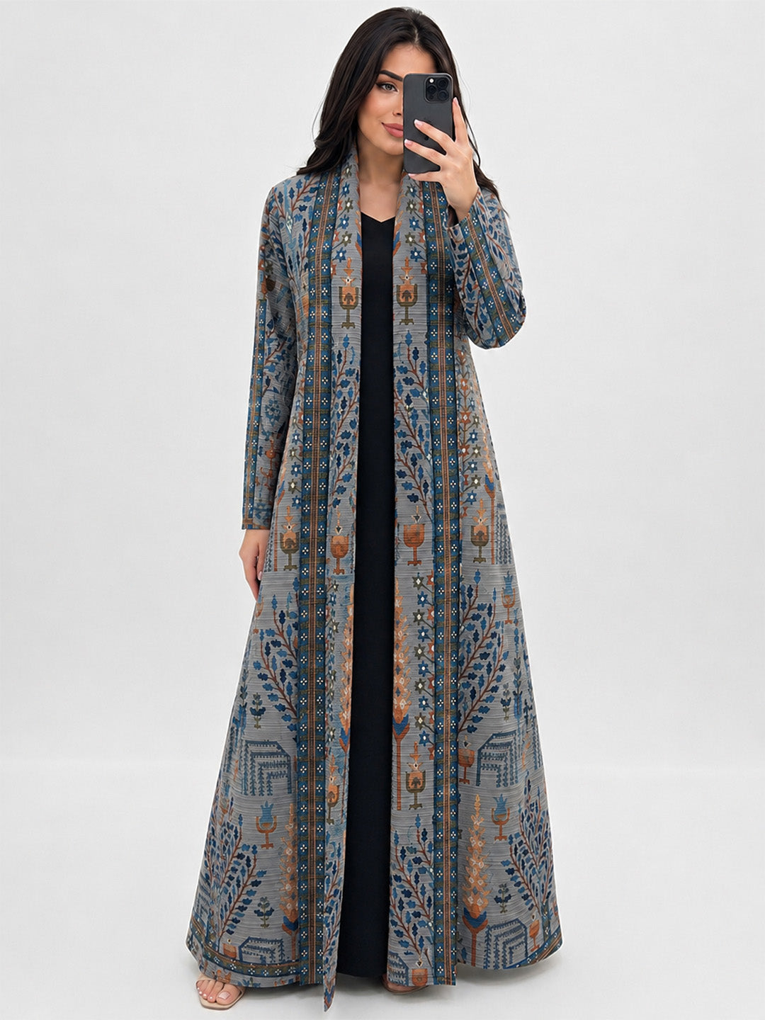 A woman takes a mirror selfie wearing a long, open-front, floor-length kaftan dress with a detailed blue, gray, and brown geometric and floral pattern over a black dress, set against a plain light gray background.