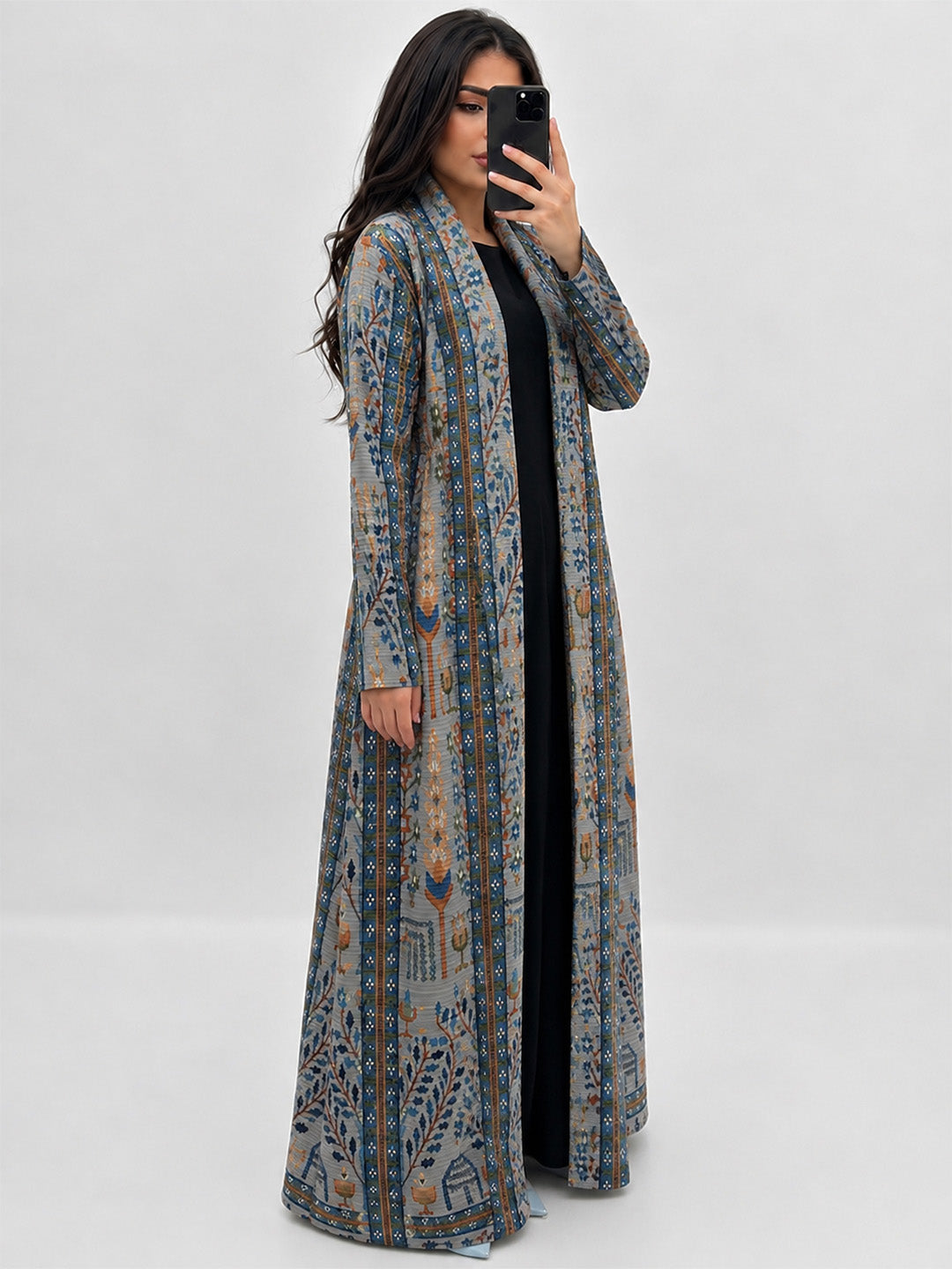 A woman modeling a long, open-front maxi cardigan with a detailed blue, gray, and brown pattern over a black dress, holding a black smartphone in a mirror selfie style against a light gray background.