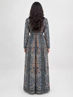 A woman viewed from the back wearing a long-sleeved maxi dress with an intricate blue, gray, and beige patterned print, set against a plain light gray studio background.