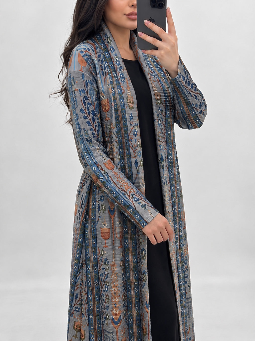A woman takes a mirror selfie wearing a long, open-front cardigan with a blue, gray, and rust-orange vertical pattern over a black dress, against a plain light gray background.
