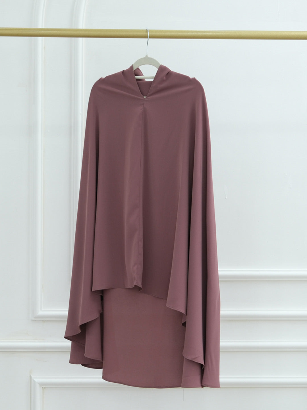 A dusty rose mauve long-sleeve draped top with a high-low hemline, hanging on a white hanger from a gold clothing rack against a white paneled wall.