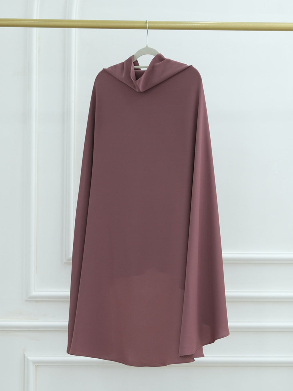 A dusty rose mauve draped cape or poncho-style dress hanging on a white hanger from a gold clothing rack against a white paneled wall.