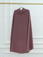 A dusty rose mauve draped cape or poncho-style dress hanging on a white hanger from a gold clothing rack against a white paneled wall.