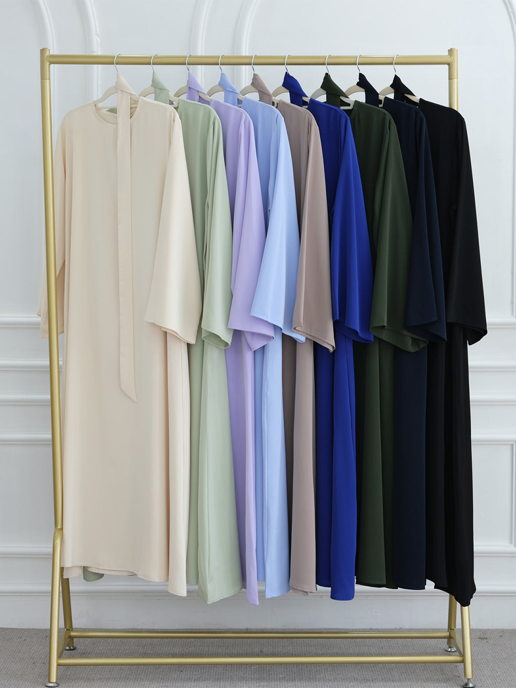 A gold clothing rack displaying a row of long, loose-fitting kaftan-style dresses in multiple colors, including beige, sage green, lavender, light blue, taupe, royal blue, forest green, and black, set against a white paneled wall.