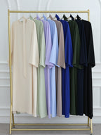 A gold clothing rack displaying a row of long, loose-fitting kaftan-style dresses in multiple colors, including beige, sage green, lavender, light blue, taupe, royal blue, forest green, and black, set against a white paneled wall.