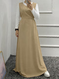A woman takes a mirror selfie wearing a long beige sleeveless maxi dress over a white long-sleeve collared shirt. She is standing on a gray carpeted floor against a white paneled wall.