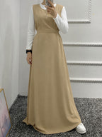 A woman takes a mirror selfie wearing a long beige sleeveless maxi dress over a white long-sleeve collared shirt. She is standing on a gray carpeted floor against a white paneled wall.